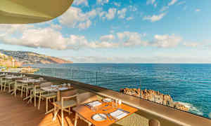 Ravintolaterassi, jossa pöytiä ja tuoleja, merelle avautuvalla ja kalliorantaisella näkymällä kirkkaan taivaan alla. (Tekoälyn luoma teksti)