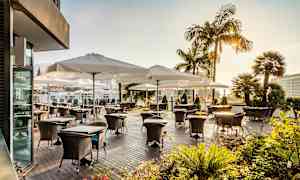 Ulkoiluterassi, jossa pöytiä ja tuoleja valkoisten aurinkovarjojen alla, ympäröitynä vihreillä kasveilla ja palmupuilla. (Tekoälyn luoma teksti)