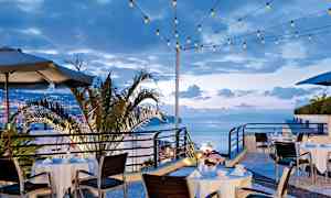 Ulkoravintola katettuine pöytineen ja tuoleineen, ympärillä valosarjoja ja näkymä kaupunkiin ja mereen. (Tekoälyn luoma teksti)