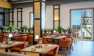 Hyvin valaistu ravintolan sisustus, jossa on pöytiä, tuoleja ja vihreitä kasveja koristeena. Taustalla suuria ikkunoita, jotka tarjoavat näkymän rakennuksesta kellotornilla. (Tekoälyn luoma teksti)