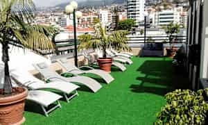 Terassi, jossa on tekonurmea, aurinkotuoleja ja ruukkukasveja. Taustalla näkyy kaupungin rakennuksia. (Tekoälyn luoma teksti)