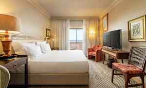 Hotellihuone, jossa on suuri sänky, yöpöytä, sohva, televisio ja kirjoituspöytä. Verhot ja lasiovet johtavat parvekkeelle. (Tekoälyn luoma teksti)