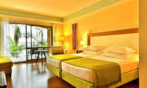 Hotellihuone, jossa on kaksi erillistä vuodetta, vihreät peitot ja istuinryhmä, näkymä ulkona oleviin palmuihin suurten lasi-ikkunoiden läpi. (Tekoälyn luoma teksti)