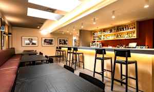 Moderni baari-alue, jossa pääosin mustia pöytiä ja tuoleja sekä hyvin valaistu baaritiski täynnä pulloja ja laseja. (Tekoälyn luoma teksti)