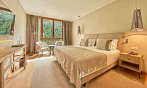 Hotellihuone, jossa on leveä sänky, kaksi nojatuolia ikkunan ääressä ja pöytä. (Tekoälyn luoma teksti)