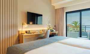 Hotellihuone, jossa työpöytä, tuoli ja televisio, sekä parveke näköalalla palmuihin ja merelle. (Tekoälyn luoma teksti)