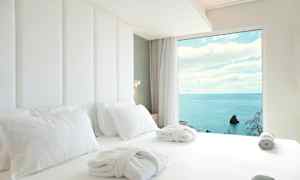 Hotellihuoneen sänky, jossa valkoiset lakanat ja kylpytakit, sekä merinäkymä ikkunasta. (Tekoälyn luoma teksti)