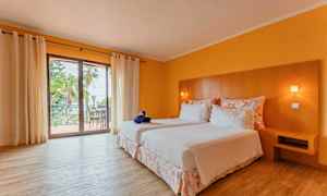 Hotellihuone, jossa on kaksi sänkyä, valkoiset vuodevaatteet, puinen pääty ja parveke näkymällä. Huoneessa on keltaiset seinät ja puulattia. (Tekoälyn luoma teksti)