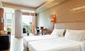 Hotellihuone, jossa kaksi yhden hengen vuodetta, istuinryhmä ja vuoristomaisema lasioven kautta. (Tekoälyn luoma teksti)