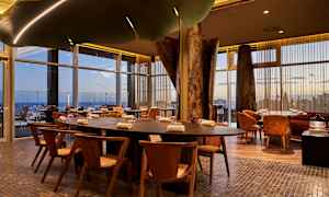 Pöytiä ja tuoleja modernissa ravintolassa, jossa on suuret lasi-ikkunat. (Tekoälyn luoma teksti)