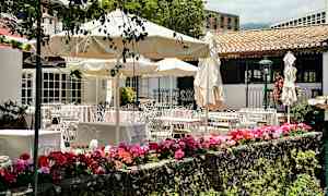 Tämä kuva esittää ulkoterassia, jossa on pöytiä, tuoleja ja aurinkovarjoja sekä värikkäitä kukkia etualalla. (Tekoälyn luoma teksti)