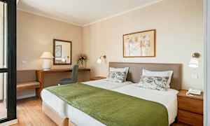 Kuva hotellihuoneesta, jossa on kaksi yksittäistä vuodetta, valkoiset vuodevaatteet ja vihreät päiväpeitot sekä taustalla työpöytä ja tuoli peilin kera. (Tekoälyn luoma teksti)