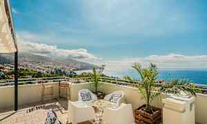 Näköala terassilta, jossa istuimia, palmuja ja panoraamanäkymä kaupunkiin, vuorille ja merelle. (Tekoälyn luoma teksti)