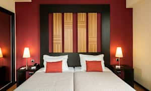 Kaksi sänkyä, joissa valkoiset vuodevaatteet ja punaiset tyynyt, taustalla puupaneeleilla varustettu punainen seinä. (Tekoälyn luoma teksti)