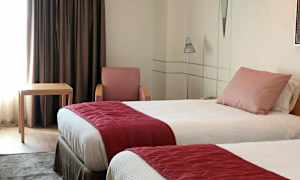 Hotellihuone, jossa kaksi yhden hengen sänkyä, punainen nojatuoli, pieni pöytä ja tummat verhot. (Tekoälyn luoma teksti)