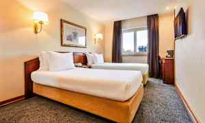 Kuva hotellihuoneesta, jossa on kaksi erillistä vuodetta, ikkuna verhoineen, seinäkoriste ja kirjoituspöytä tuoleineen. (Tekoälyn luoma teksti)