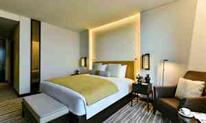 Nykyaikainen hotellihuone, jossa on parivuode, kaksi yöpöytää ja mukava nojatuoli. (Tekoälyn luoma teksti)