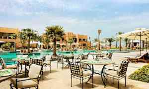 Ulkoravintola uima-altaan äärellä puutarhamaisessa ympäristössä, palmujen ja rakennusten ympäröimänä. (Tekoälyn luoma teksti)