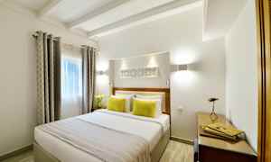 Vaalea makuuhuone, jossa on parisänky ja kaksi keltaista tyynyä sekä päiväpeite. Ikkunassa on verhot, ja huoneessa on työpiste, jossa on pöytä ja lamppu. (Tekoälyn luoma teksti)