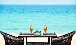 Kaksi rottinkituolia pöydän ympärillä, ja pöydällä on kaksi lasillista mehua ja kulho tuoreita hedelmiä trooppisella rannalla. (Tekoälyn luoma teksti)