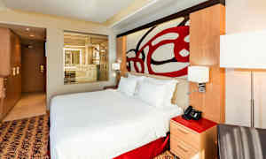 Moderni hotellihuone, jossa on suuri sänky ja läpinäkyvä lasiseinä kylpyhuoneeseen. (Tekoälyn luoma teksti)