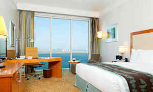 Moderni hotellihuone, jossa on suuri sänky, työpöytä ja näkymä suurista ikkunoista merelle ja taustalla näkyvään kaupunkiin. (Tekoälyn luoma teksti)