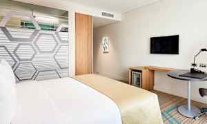 Hotellihuone, jossa on valkoisiin ja beigeihin väreihin päällystetty sänky. Huoneessa on myös työpöytä, seinälle kiinnitetty televisio ja minijääkaappi. (Tekoälyn luoma teksti)