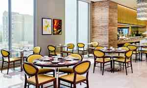 Kuva valoisasta ja modernista ravintolasta, jossa on pyöreitä pöytiä, keltaisia tuoleja ja hyvin varusteltu buffet-alue. (Tekoälyn luoma teksti)