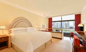 Hotellihuone, jossa on parivuode, yöpöydät ja suuret ikkunat, joista on näkymä kaupunkimaisemaan. (Tekoälyn luoma teksti)