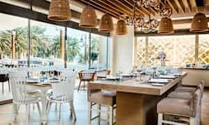 Ravintolan sisätilat, joissa pöydät ovat katettu posliinilla ja lasilla sekä rottinkivalaisimet roikkuvat katosta. Ulkona näkyy palmupuita. (Tekoälyn luoma teksti)