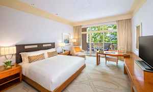 Hotellihuone, jossa on suuri sänky, kirjoituspöytä ja parveke näkymällä vehreään ympäristöön. (Tekoälyn luoma teksti)