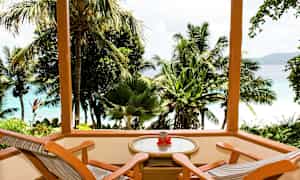 Puuterassi, jossa on kaksi tuolia ja pöytä, jolta avautuu näkymä trooppisiin palmuihin ja siniseen mereen. (Tekoälyn luoma teksti)