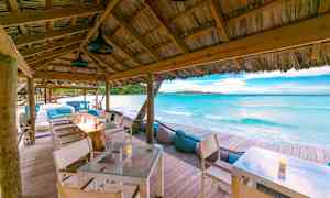 Rannalla ulkotila, jossa on pöytiä puukatteen alla lähistöllä kirkasta sinistä merta ja hiekkaa. (Tekoälyn luoma teksti)