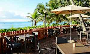 Meren äärellä sijaitseva ulkoterassi, jossa on pöytiä, tuoleja ja aurinkovarjoja, ympärillä vehreää kasvillisuutta ja palmupuita. (Tekoälyn luoma teksti)