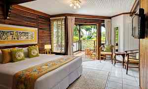 Makuuhuone, jossa on parivuode ja värikkäillä tekstiileillä sekä seinäkoristeilla sisustettu, puupintaiset seinät ja suuri ikkuna, josta näkymä vehreälle verannalle. (Tekoälyn luoma teksti)