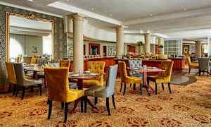 Tyylikäs ruokailutila, jossa on useita katettuja pöytiä, mukavat sinapinkeltaiset ja harmaat tuolit sekä koristeellisia pylväitä ja peilejä. (Tekoälyn luoma teksti)