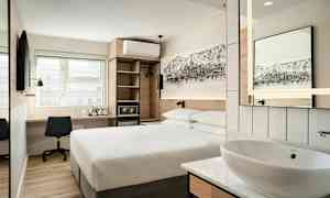 Moderni hotellihuone, jossa on parivuode, kirjoituspöytä ja tuoli, seinälle asennettu televisio sekä avonaulakko ja pieni pesuallas peileineen. (Tekoälyn luoma teksti)