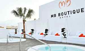 Hotellialue, jossa on valkoisia aurinkotuoleja ja oransseja pyyhkeitä altaan äärellä, palmu ja valkoiset seinät. (Tekoälyn luoma teksti)