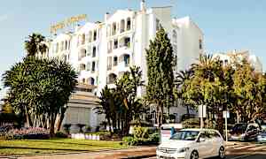 Valkoinen rakennus hotellikyltillä, ympäröitynä vehreydellä ja pysäköidyillä autoilla. (Tekoälyn luoma teksti)