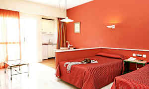 Hotellihuone, jossa on kaksi erillistä sänkyä, punaiset vuodevaatteet ja punainen seinä sekä pieni keittiö ja lasipöytä taustalla. (Tekoälyn luoma teksti)