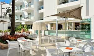 Ulkotila, jossa on pöytiä, tuoleja ja aurinkovarjoja rakennuksen edessä. (Tekoälyn luoma teksti)