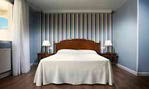 Hotellihuone, jossa sänky keskellä, kaksi yöpöytää lampuilla ja siniraitainen tapetti. (Tekoälyn luoma teksti)