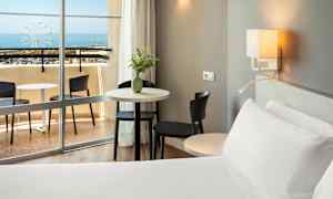 Hotellihuone, jossa vuode, pöytä ja tuolit lähellä parveketta merinäköalalla. (Tekoälyn luoma teksti)