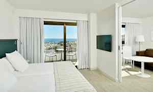 Hotellihuoneen sisätilat, joissa on parivuode, erillinen istuma-alue ja parveke, jolta on näkymä merelle ja satamaan. (Tekoälyn luoma teksti)