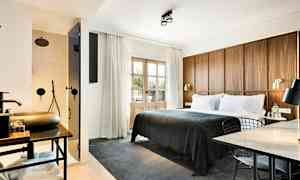 Hotellihuone, jossa iso sänky tummanharmaalla peitteellä, puinen paneelipääty, työtila pöydällä ja tuolilla sekä näkyvissä oleva kylpyhuone, jossa suihku lasioven takana. (Tekoälyn luoma teksti)