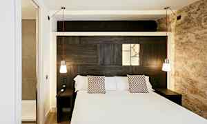 Hotellihuone, jossa on parivuode, kaksi roikkuvaa lamppua ja valopaneeli sängyn takana puuseinässä. (Tekoälyn luoma teksti)