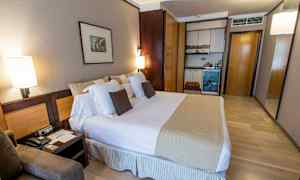 Hotellihuone, jossa on iso sänky, tyynyjä, yöpöytä ja pieni keittiöosa kaappeineen. (Tekoälyn luoma teksti)