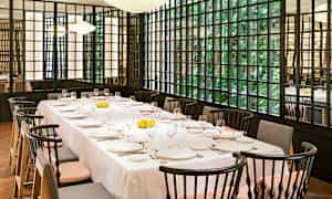 Kattaus ravintolassa, jossa on tyylikkäitä tuoleja ja suuret ikkunat, joiden takana on vihreyttä. (Tekoälyn luoma teksti)