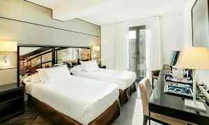 Hotellihuone, jossa kaksi yhden hengen vuodetta, kirjoituspöytä ja tuoli sekä parveke valkoisilla verhoilla. (Tekoälyn luoma teksti)