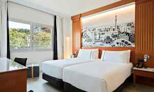 Hotellihuone, jossa on kaksi erillistä sänkyä, kirjoituspöytä, näkymä ikkunasta vihreään ympäristöön ja seinällä suuri mustavalkoinen kuva koristeena. (Tekoälyn luoma teksti)